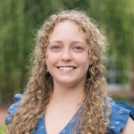 A woman with curly blonde hair and a blue top smiles outdoors, with green foliage blurred in the background.