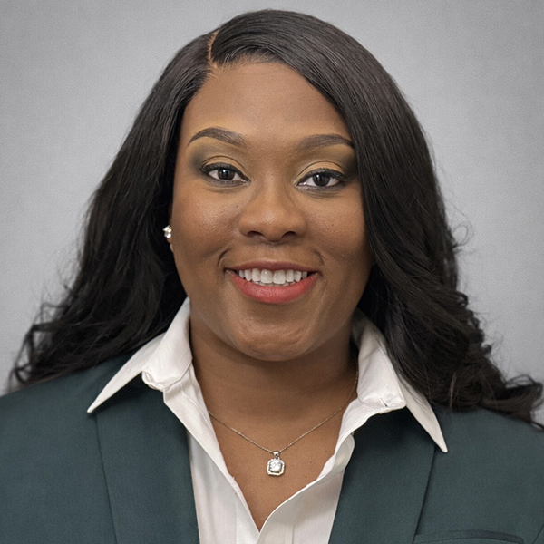 Woman with long dark hair wearing a green blazer, white collared shirt, and a necklace, smiling against a plain gray background.