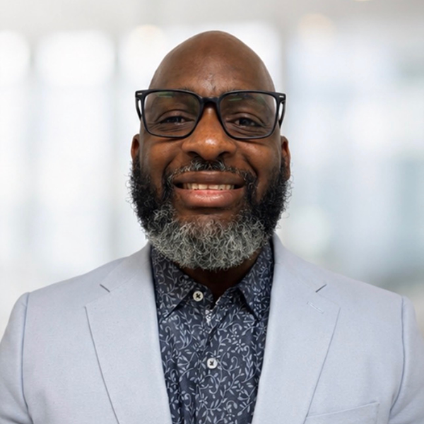 A man with a gray beard and glasses, wearing a light-colored blazer and a patterned shirt, smiles at the camera with a blurred indoor background.