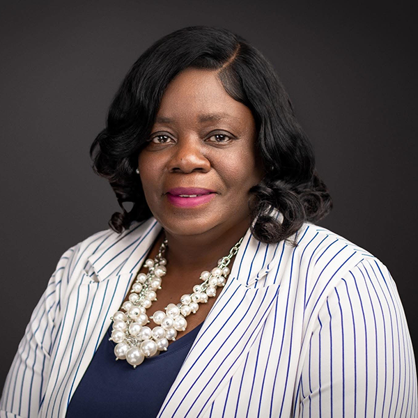Woman with shoulder-length black hair wearing a white pinstripe blazer, navy top, and pearl necklace, posed against a dark background.