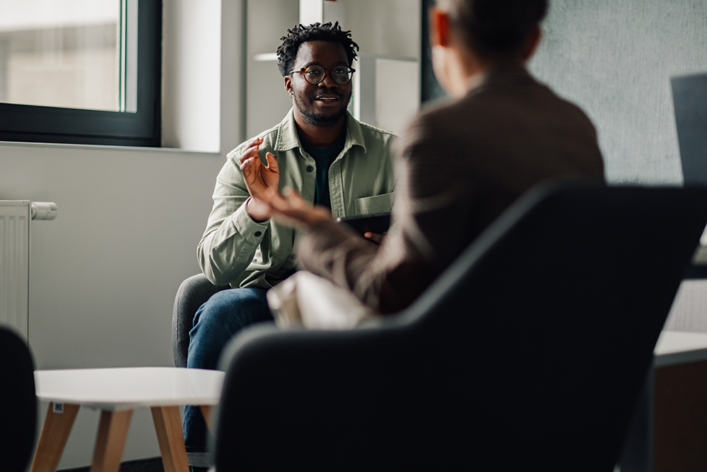 Two people sit across from each other in armchairs, having a discussion in a modern office setting. One person gestures with their hand while listening intently.